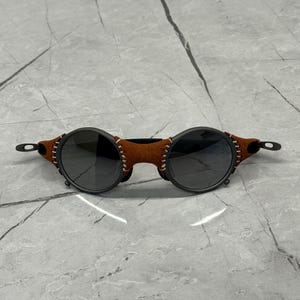 May include: Round sunglasses with dark lenses and a black frame. The temples are adorned with brown leather and white stitching. The sunglasses have a unique, vintage-inspired design.