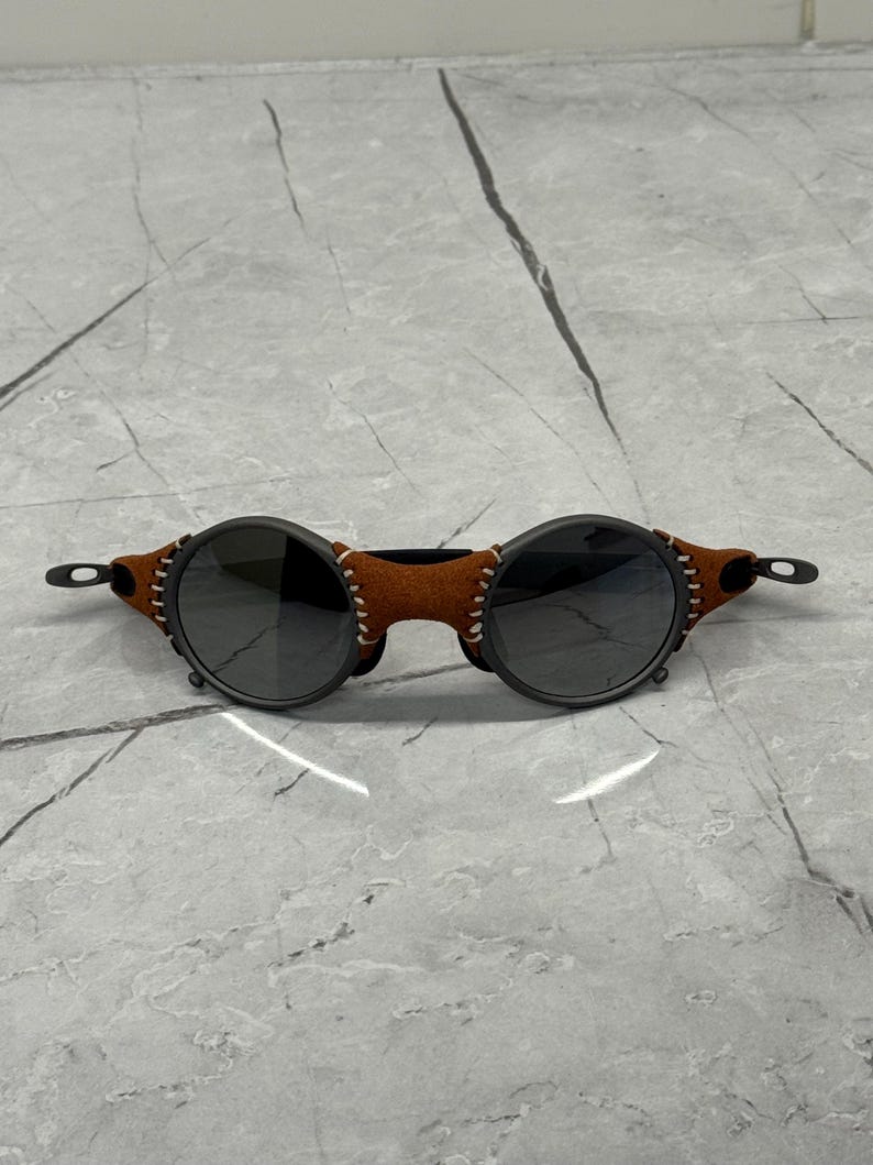 May include: Round sunglasses with dark lenses and a black frame. The temples are adorned with brown leather and white stitching. The sunglasses have a unique, vintage-inspired design.