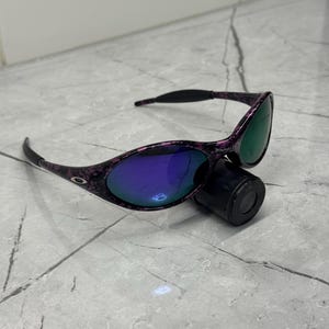 May include: Purple and black patterned sunglasses with mirrored lenses reflecting blue and green. The sunglasses are resting on a black cylindrical object. The frame has a dark pattern.