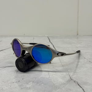 Może przedstawiać: Srebrne okulary przeciwsłoneczne z niebieskimi soczewkami. Okrągłe soczewki odbijają żywy niebieski odcień. Okulary przeciwsłoneczne spoczywają na czarnym cylindrycznym obiekcie na jasnym tle.