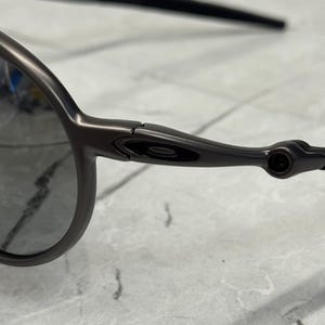 May include: Close-up of dark grey sunglasses. The sunglasses have a sleek, modern design with a dark frame and lenses. The arms of the sunglasses are thin and curve slightly. The lenses are a dark grey colour.