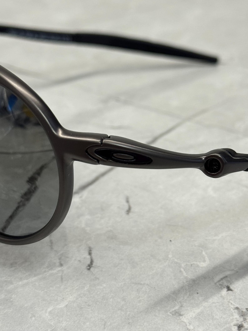 May include: Close-up of dark grey sunglasses. The sunglasses have a sleek, modern design with a dark frame and lenses. The arms of the sunglasses are thin and curve slightly. The lenses are a dark grey colour.