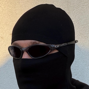 May include: A person wearing a black balaclava and dark-tinted sunglasses. The sunglasses have a clear frame and dark lenses. The balaclava covers the head and neck, leaving only the eyes visible.