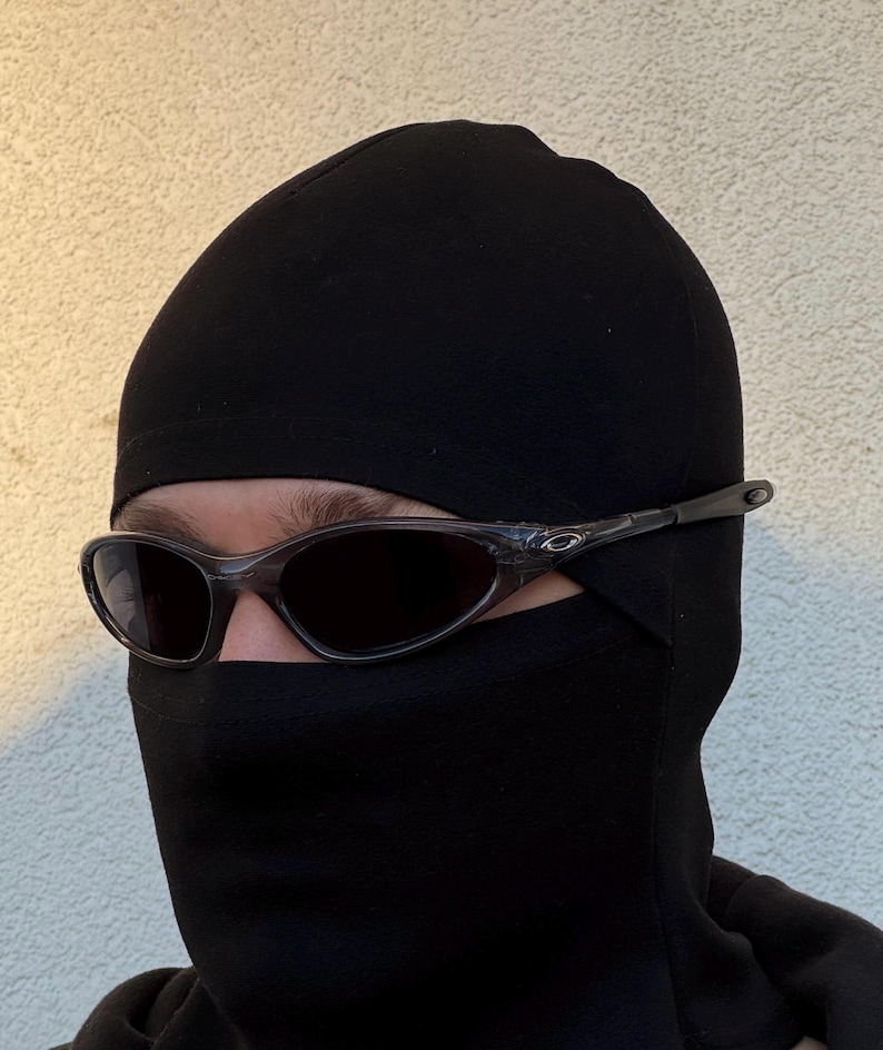 May include: A person wearing a black balaclava and dark-tinted sunglasses. The sunglasses have a clear frame and dark lenses. The balaclava covers the head and neck, leaving only the eyes visible.