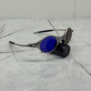May include: Silver-framed sunglasses with blue and brown lenses. The sunglasses have a sleek, modern design with curved temples and a small, black cylindrical case. The lenses are oval-shaped and reflect light.