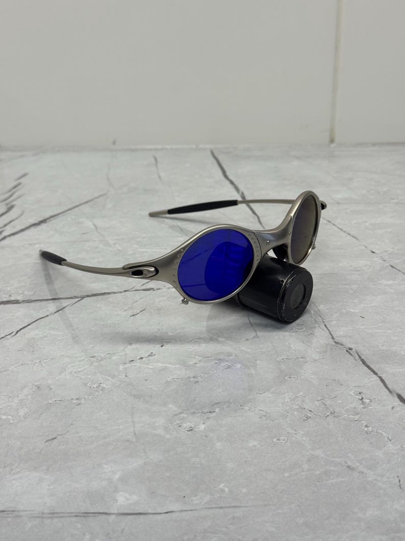 May include: Silver-framed sunglasses with blue and brown lenses. The sunglasses have a sleek, modern design with curved temples and a small, black cylindrical case. The lenses are oval-shaped and reflect light.
