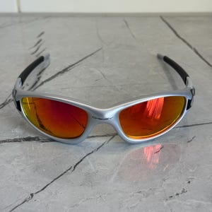 May include: Silver-framed sunglasses with red-orange lenses. The sunglasses have a curved design with black arms and the brand name "Oakley" visible on the frame. The lenses reflect the surrounding environment.