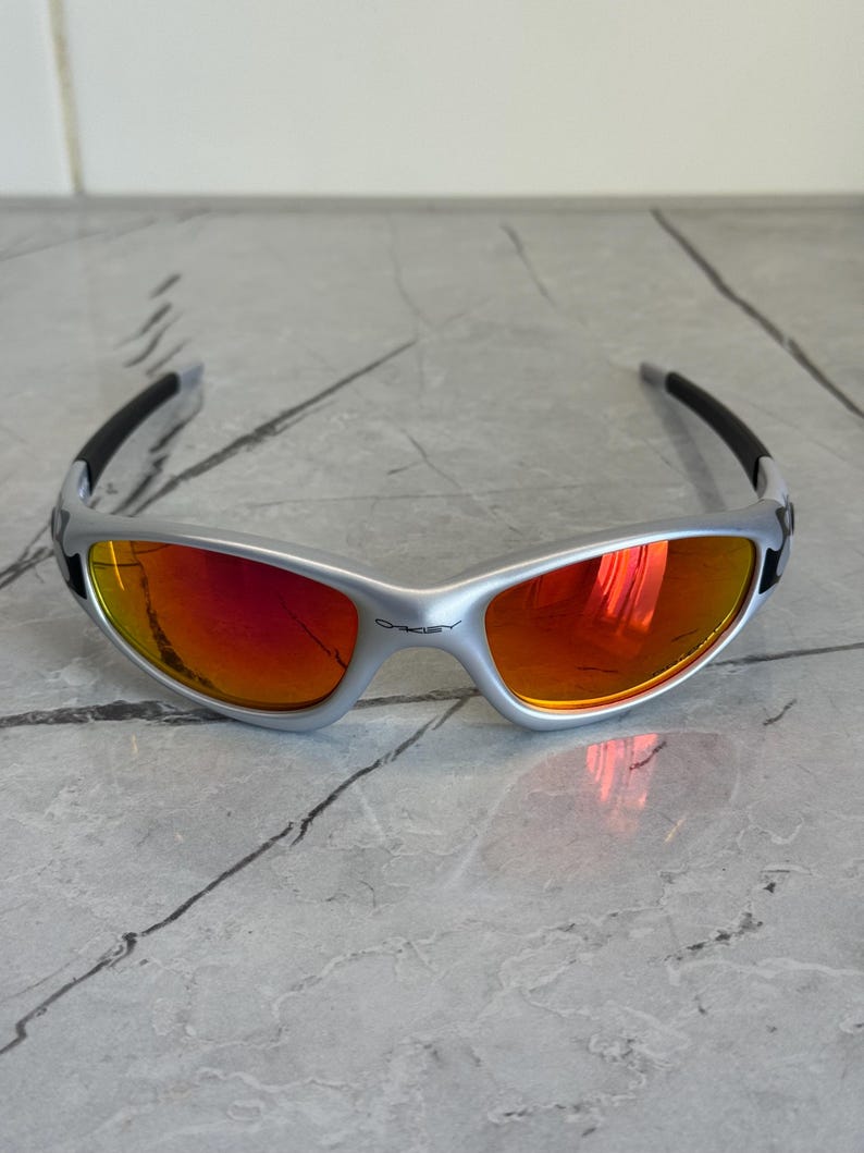 May include: Silver-framed sunglasses with red-orange lenses. The sunglasses have a curved design with black arms and the brand name "Oakley" visible on the frame. The lenses reflect the surrounding environment.