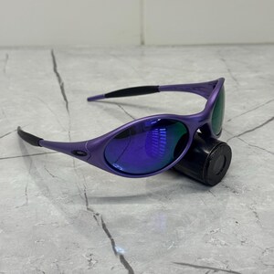 May include: Purple sunglasses with a sleek, sporty design. The frames are a vibrant purple, complemented by dark lenses that reflect a blue hue. The temples are black, and the glasses rest on a black cylindrical object.