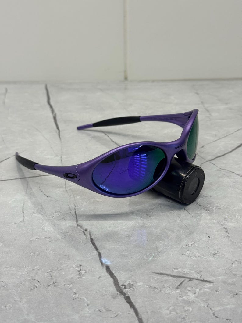 May include: Purple sunglasses with a sleek, sporty design. The frames are a vibrant purple, complemented by dark lenses that reflect a blue hue. The temples are black, and the glasses rest on a black cylindrical object.