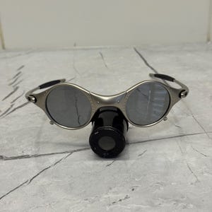 May include: Silver-framed sunglasses with round lenses and a black nose bridge. The sunglasses have a unique design with a small, cylindrical attachment below the lenses. The lenses are reflective, and the frame is metallic.