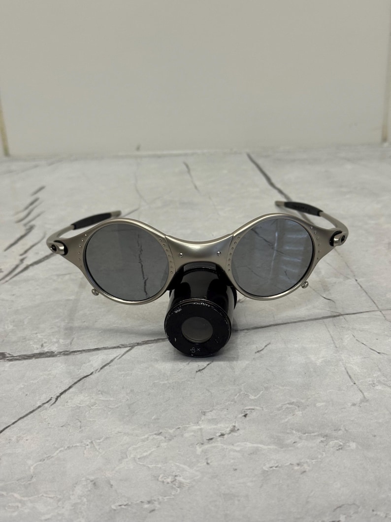 May include: Silver-framed sunglasses with round lenses and a black nose bridge. The sunglasses have a unique design with a small, cylindrical attachment below the lenses. The lenses are reflective, and the frame is metallic.