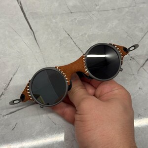 May include: Round sunglasses with dark lenses and a brown leather bridge. The leather is stitched with white thread. The frames are a dark gray color. The sunglasses have a unique, vintage style.