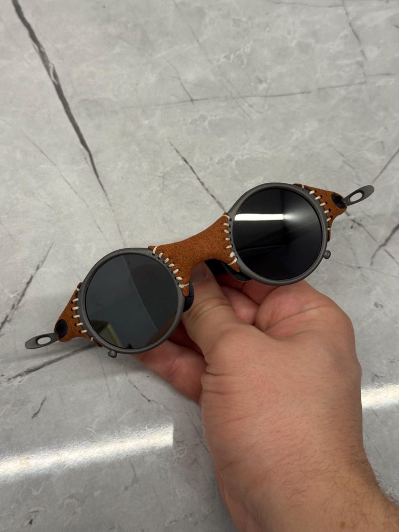 May include: Round sunglasses with dark lenses and a brown leather bridge. The leather is stitched with white thread. The frames are a dark gray color. The sunglasses have a unique, vintage style.