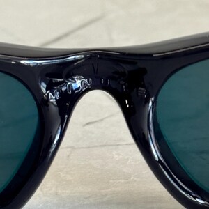 May include: Close-up of black sunglasses with green lenses. The sunglasses have a curved design and a small logo on the bridge. The lenses are a dark green color, and the frames are glossy black.