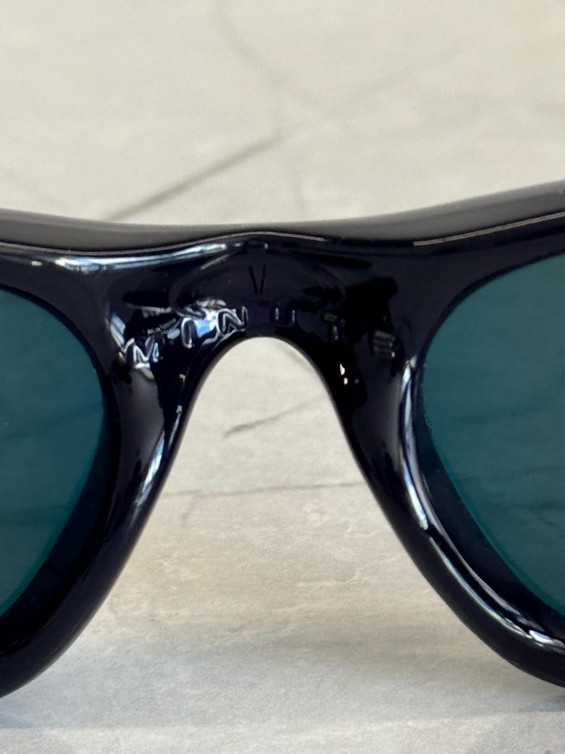 May include: Close-up of black sunglasses with green lenses. The sunglasses have a curved design and a small logo on the bridge. The lenses are a dark green color, and the frames are glossy black.