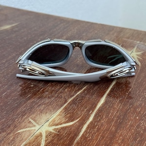 Puede incluir: Un par de gafas de sol plateadas y grises con lentes oscuras. Las gafas de sol tienen un dise&ntilde;o moderno con una montura met&aacute;lica y est&aacute;n sobre una superficie de madera.