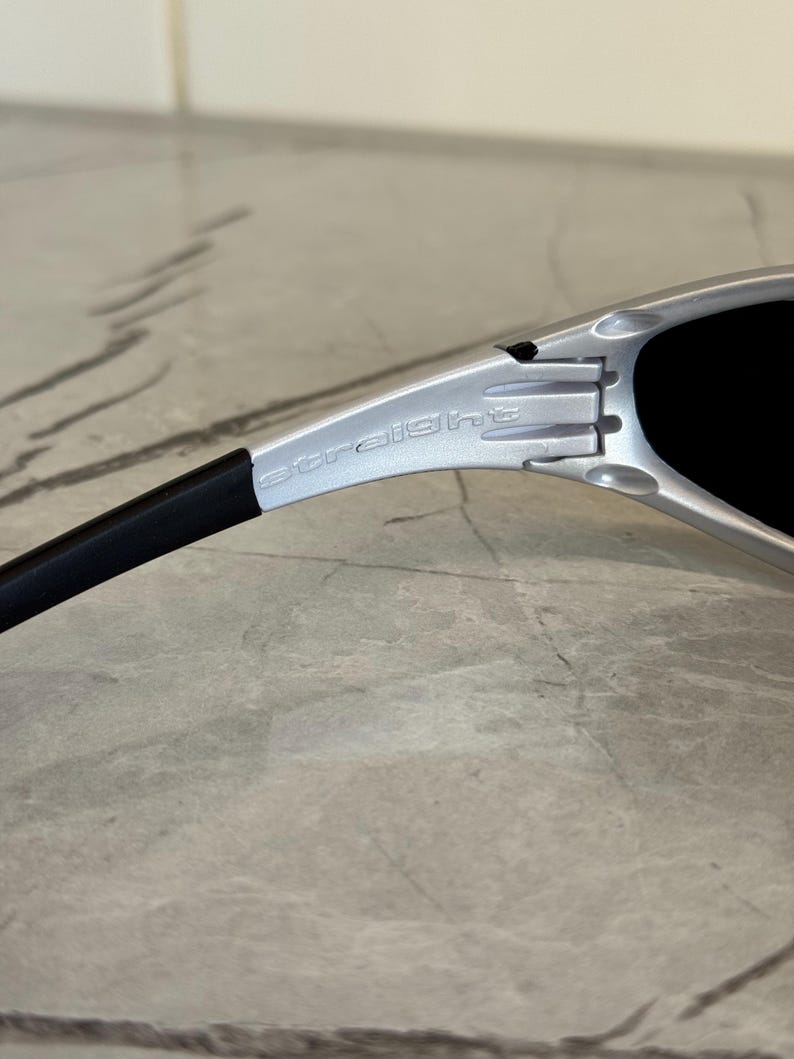 May include: Close-up of silver sunglasses with black lenses. The word "straight" is printed on the silver frame. The sunglasses have a black arm.