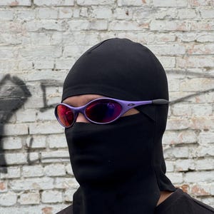 May include: A person wearing purple-framed sunglasses with red lenses and a black balaclava. The background is a weathered white brick wall. The sunglasses have a sporty design.