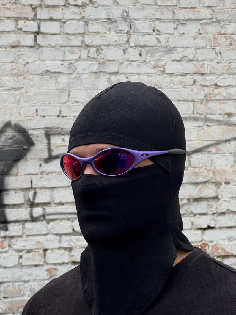 May include: A person wearing purple-framed sunglasses with red lenses and a black balaclava. The background is a weathered white brick wall. The sunglasses have a sporty design.