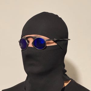 以下が含まれることがあります： 青いレンズの丸いシルバーフレームのサングラスが、黒いバラクラバの上からかけられています。バラクラバは頭と首を覆い、目だけが見えています。背景は無地のオフホワイトの壁です。
