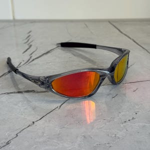 K&ouml;nnte beinhalten: Grau gerahmte Sonnenbrille mit roten und gelben Gl&auml;sern. Die Sonnenbrille hat ein schlankes, modernes Design mit einem geschwungenen Rahmen und dunklen B&uuml;geln. Die Gl&auml;ser reflektieren einen Farbverlauf.