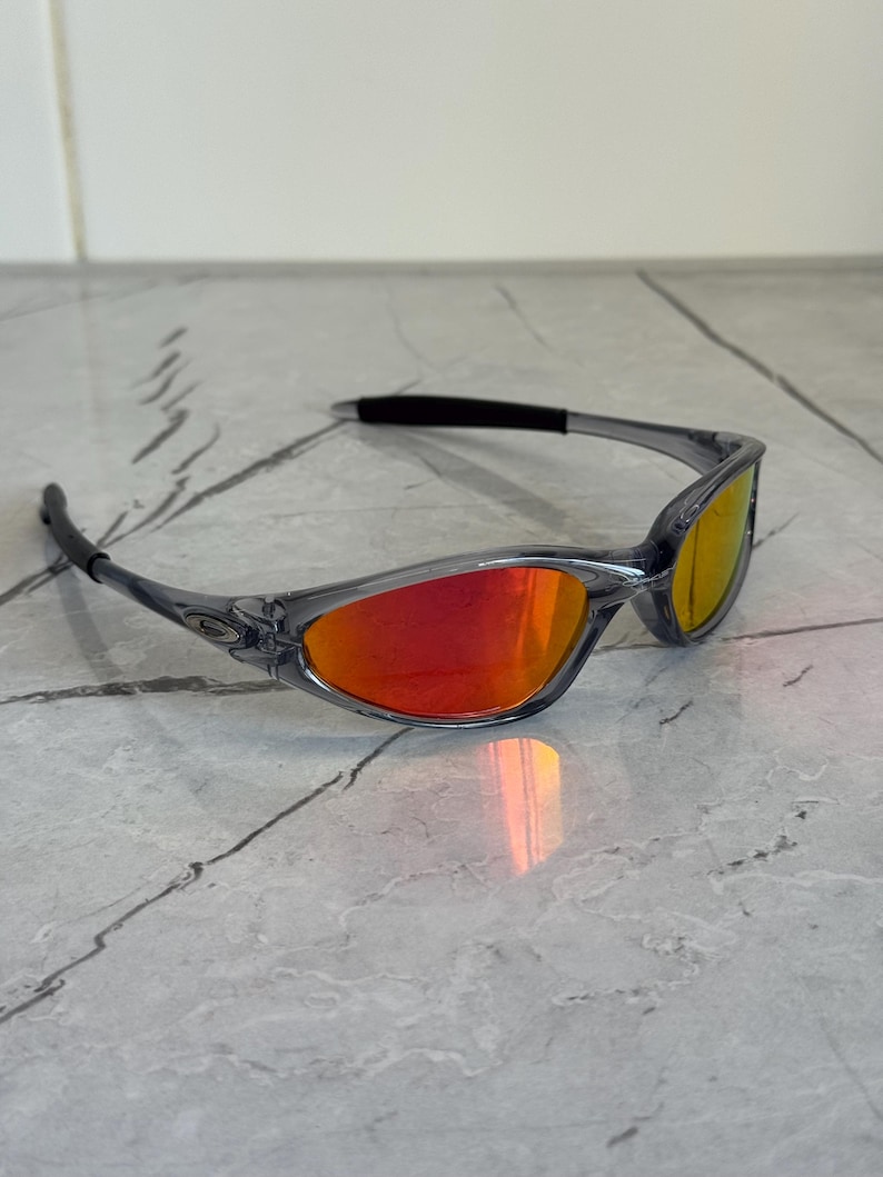 K&ouml;nnte beinhalten: Grau gerahmte Sonnenbrille mit roten und gelben Gl&auml;sern. Die Sonnenbrille hat ein schlankes, modernes Design mit einem geschwungenen Rahmen und dunklen B&uuml;geln. Die Gl&auml;ser reflektieren einen Farbverlauf.