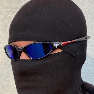 May include: Black sunglasses with blue lenses. The frames are black with a white star and red accents. The sunglasses are worn with a black balaclava.