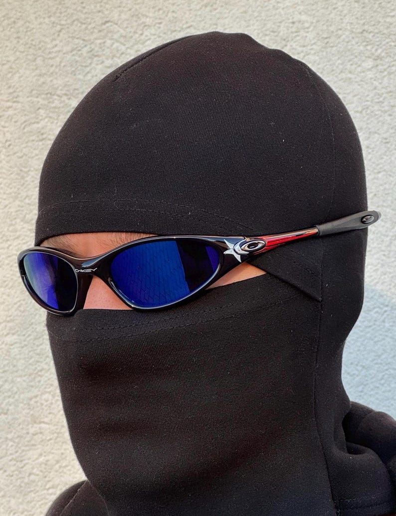 May include: Black sunglasses with blue lenses. The frames are black with a white star and red accents. The sunglasses are worn with a black balaclava.