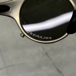 May include: Close-up of a pair of sunglasses with a metallic frame and dark lenses. The text "POLARIZED" is visible on the frame. The sunglasses have a rounded shape and a sleek, modern design.