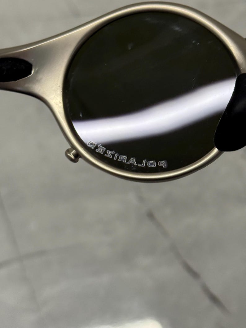 May include: Close-up of a pair of sunglasses with a metallic frame and dark lenses. The text "POLARIZED" is visible on the frame. The sunglasses have a rounded shape and a sleek, modern design.