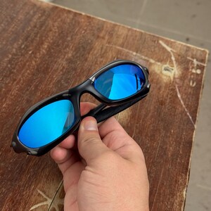 Op de afbeelding: Zwarte zonnebril met blauw gespiegelde glazen. De zonnebril heeft een strak, modern design met een gebogen montuur. De glazen reflecteren een heldere blauwe kleur, wat zowel stijl als bescherming tegen de zon biedt.