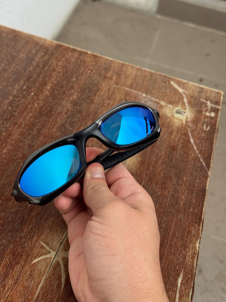 Op de afbeelding: Zwarte zonnebril met blauw gespiegelde glazen. De zonnebril heeft een strak, modern design met een gebogen montuur. De glazen reflecteren een heldere blauwe kleur, wat zowel stijl als bescherming tegen de zon biedt.