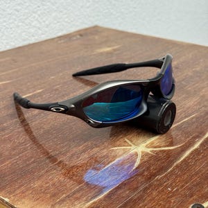 Op de afbeelding: Een zwarte zonnebril met blauw gespiegelde glazen. De zonnebril heeft een slank, modern ontwerp met een gebogen montuur en rust op een zwart cilindrisch object. De glazen reflecteren een heldere blauwe tint.