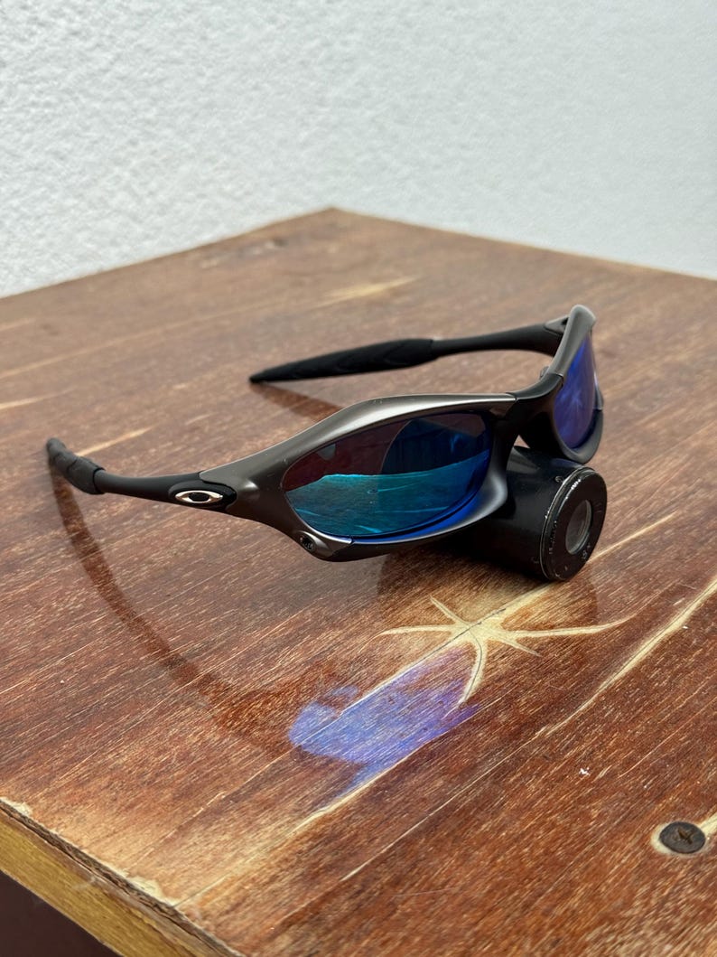 Op de afbeelding: Een zwarte zonnebril met blauw gespiegelde glazen. De zonnebril heeft een slank, modern ontwerp met een gebogen montuur en rust op een zwart cilindrisch object. De glazen reflecteren een heldere blauwe tint.