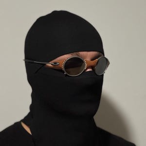 May include: Black balaclava with round, tinted sunglasses. The sunglasses have a silver frame and brown leather accents. The balaclava covers the head and neck, with only the eyes visible.