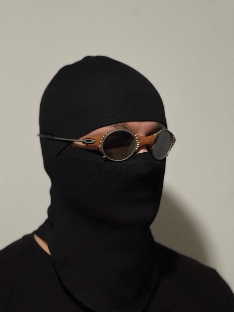 May include: Black balaclava with round, tinted sunglasses. The sunglasses have a silver frame and brown leather accents. The balaclava covers the head and neck, with only the eyes visible.