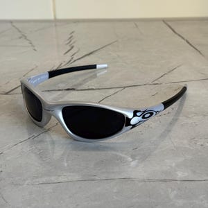 May include: Silver sunglasses with black lenses. The frames have a sleek, curved design with black temples and white accents. A decorative graphic is on the temple. The sunglasses are on a gray surface.