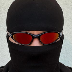 May include: Black Oakley sunglasses with red lenses are worn with a black balaclava. The sunglasses have a black frame with a white star design on the temples. The Oakley logo is visible on the frame.