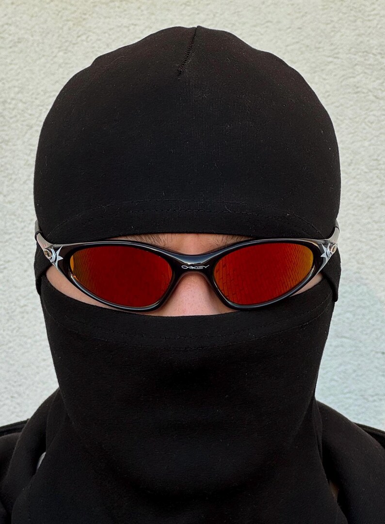 May include: Black Oakley sunglasses with red lenses are worn with a black balaclava. The sunglasses have a black frame with a white star design on the temples. The Oakley logo is visible on the frame.