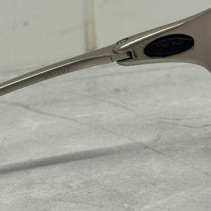 May include: Close-up of silver-toned sunglasses. The frame is a sleek, modern design with a hinge detail. The arm of the glasses has the number "04-107" printed on it. The lens area has a black oval detail.