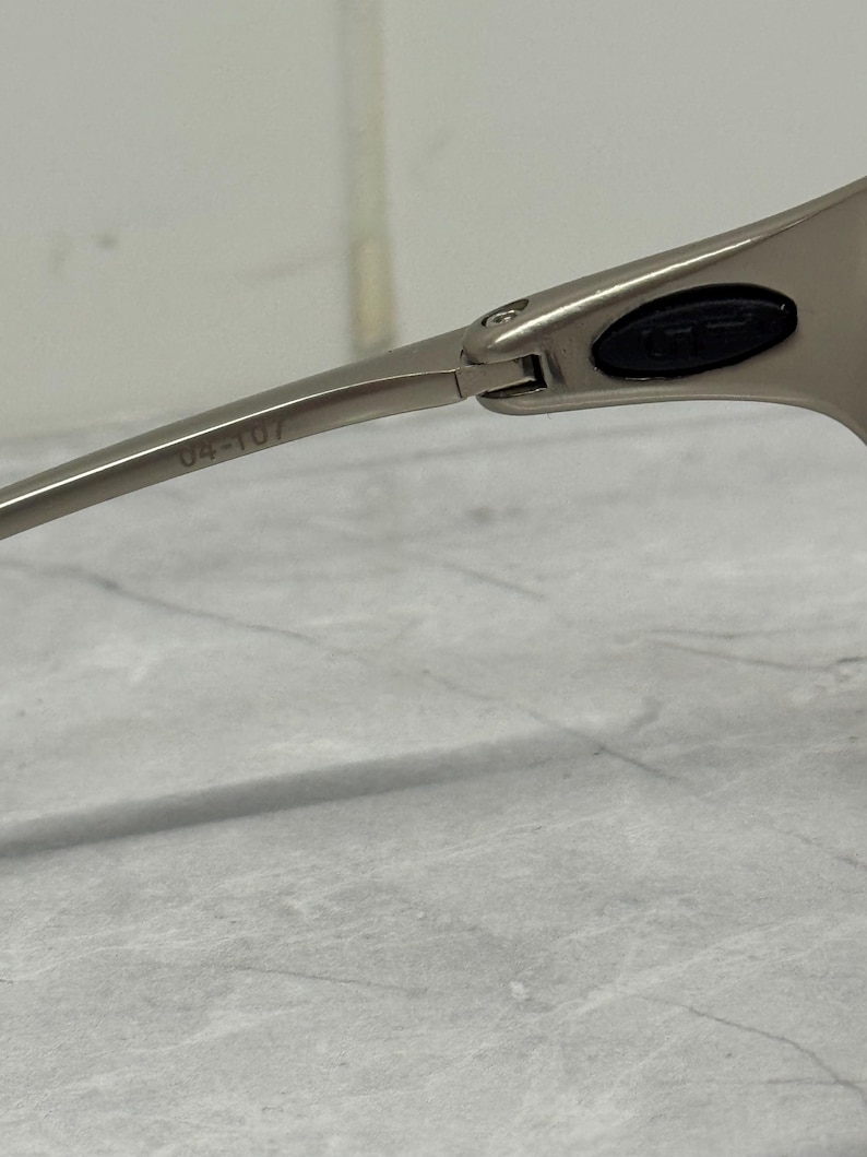 May include: Close-up of silver-toned sunglasses. The frame is a sleek, modern design with a hinge detail. The arm of the glasses has the number "04-107" printed on it. The lens area has a black oval detail.
