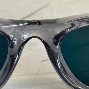 K&ouml;nnte beinhalten: Nahaufnahme einer grauen Sonnenbrille mit dunkelgr&uuml;nen Gl&auml;sern. Der Rahmen hat ein geschwungenes Design und der Markenname ist auf der Oberseite des Rahmens sichtbar. Die Gl&auml;ser sind tiefgr&uuml;n.