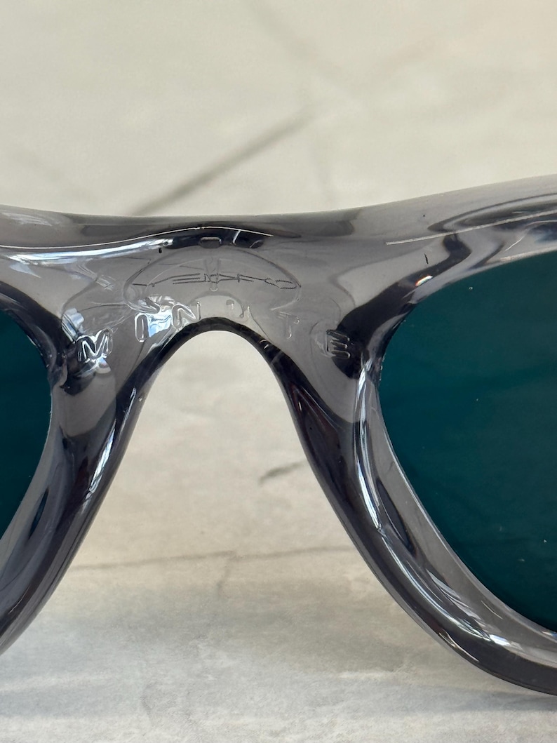 K&ouml;nnte beinhalten: Nahaufnahme einer grauen Sonnenbrille mit dunkelgr&uuml;nen Gl&auml;sern. Der Rahmen hat ein geschwungenes Design und der Markenname ist auf der Oberseite des Rahmens sichtbar. Die Gl&auml;ser sind tiefgr&uuml;n.