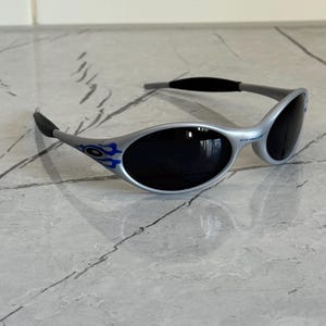 Puede incluir: Gafas de sol plateadas con lentes oscuras. Las gafas tienen un dise&ntilde;o curvo con detalles azules. Las patillas son grises con puntas negras. Las lentes son ovaladas. La montura mide unos 14 cm de ancho.