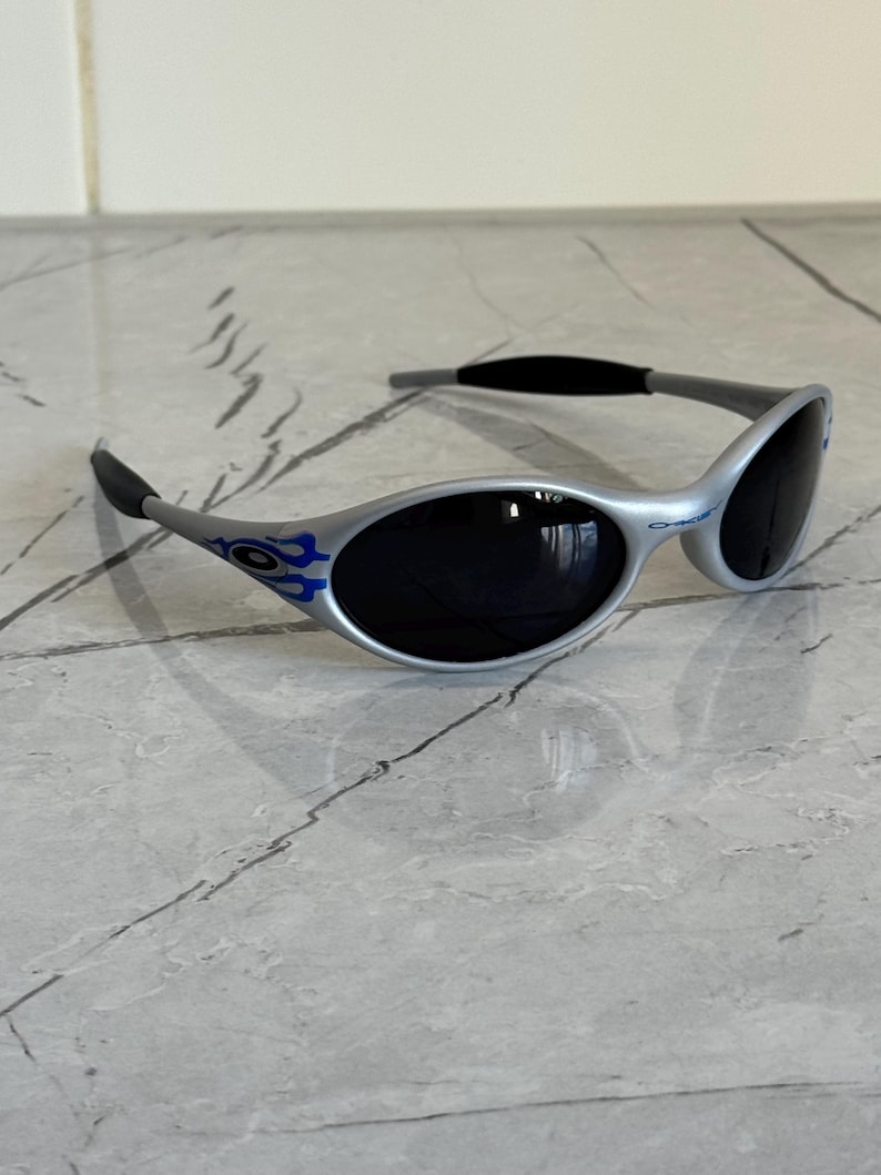Puede incluir: Gafas de sol plateadas con lentes oscuras. Las gafas tienen un dise&ntilde;o curvo con detalles azules. Las patillas son grises con puntas negras. Las lentes son ovaladas. La montura mide unos 14 cm de ancho.