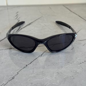 K&ouml;nnte beinhalten: Schwarze Sonnenbrille mit einem schlanken, sportlichen Design. Die Sonnenbrille hat einen gl&auml;nzend schwarzen Rahmen und dunkle Gl&auml;ser. Die B&uuml;gel sind um die Ohren gebogen. Das Oakley-Logo ist auf dem Rahmen sichtbar.