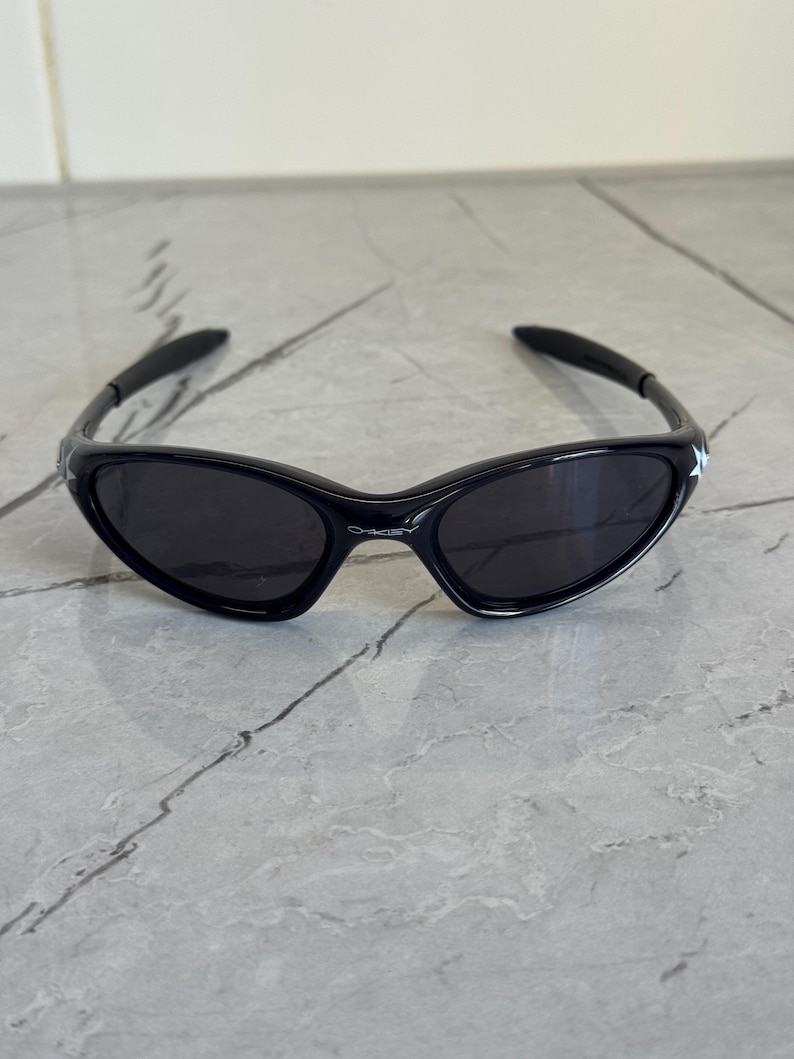 K&ouml;nnte beinhalten: Schwarze Sonnenbrille mit einem schlanken, sportlichen Design. Die Sonnenbrille hat einen gl&auml;nzend schwarzen Rahmen und dunkle Gl&auml;ser. Die B&uuml;gel sind um die Ohren gebogen. Das Oakley-Logo ist auf dem Rahmen sichtbar.