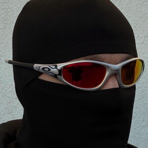 May include: Silver-framed sunglasses with red lenses are worn over a black balaclava. The sunglasses have a decorative design on the temples. The lenses reflect the surrounding environment. The balaclava covers the head and neck.