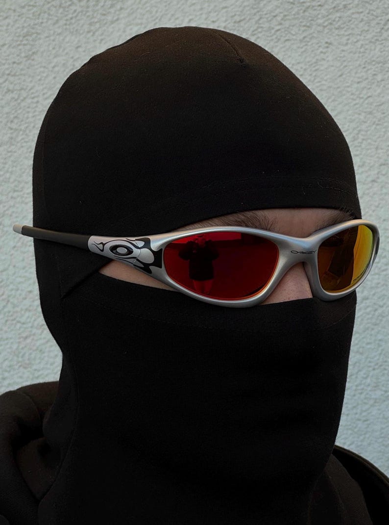 May include: Silver-framed sunglasses with red lenses are worn over a black balaclava. The sunglasses have a decorative design on the temples. The lenses reflect the surrounding environment. The balaclava covers the head and neck.